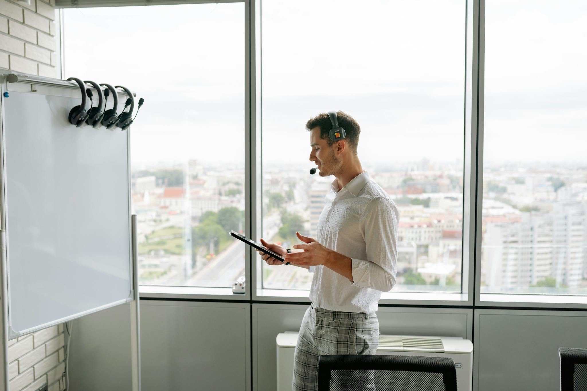 Man with headset and tablet in office by a whiteboard and large windows overlooking cityscape.